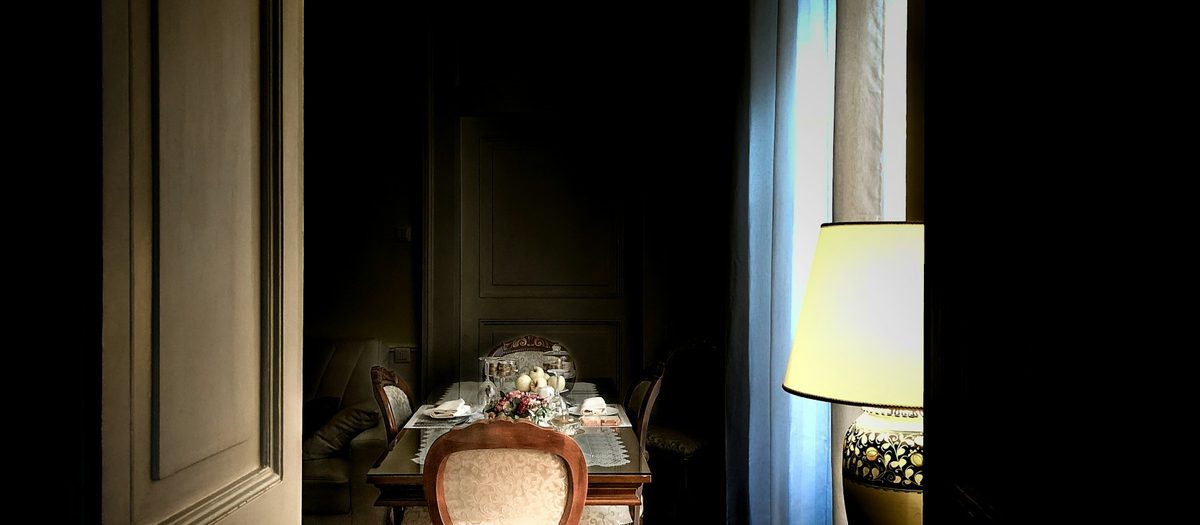 Een schemerige kamer vanuit de deur gezien, met de lamp aan en de tafel gedekt · Italiaanse grammatica A1