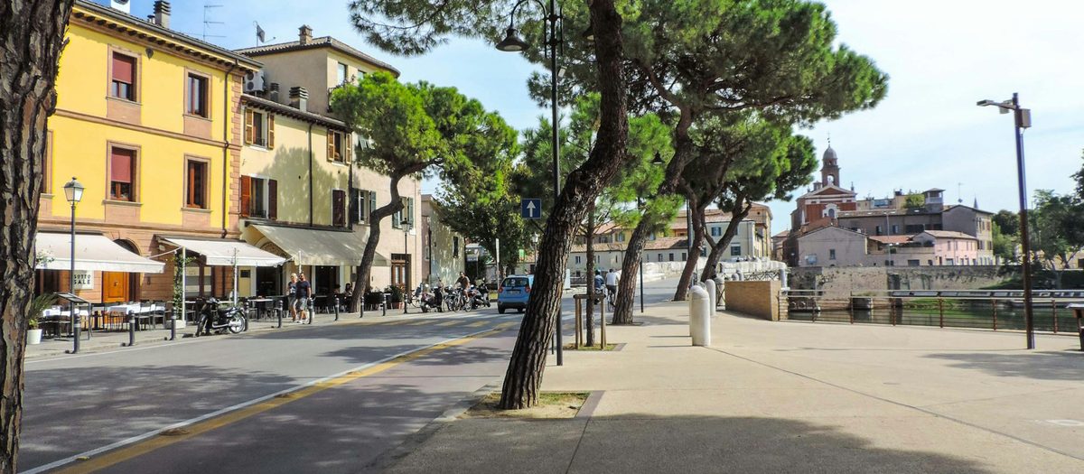 Een lommerrijke straat langs het water, met tafeltjes onder de luifels en een klokkentoren · Italiaanse grammatica C1