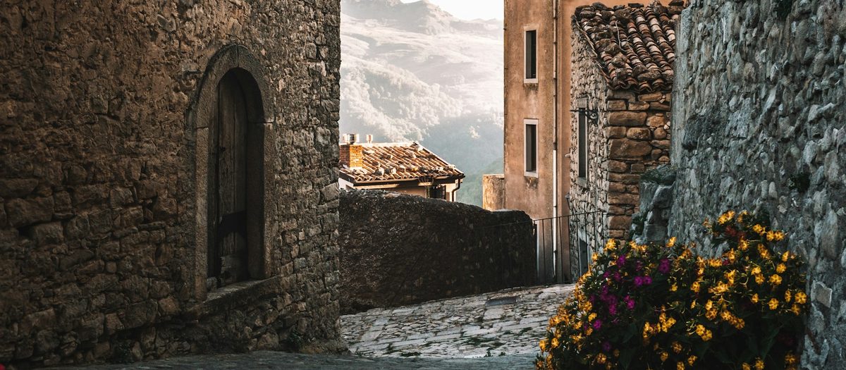 Een stenen steegje in een oud dorp, met een boog en gele bloemen · Italiaanse grammatica A1