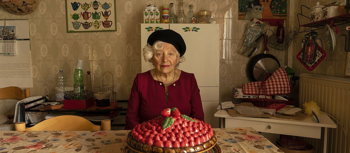 Een oudere vrouw in haar keuken, met een aardbeientaart op tafel