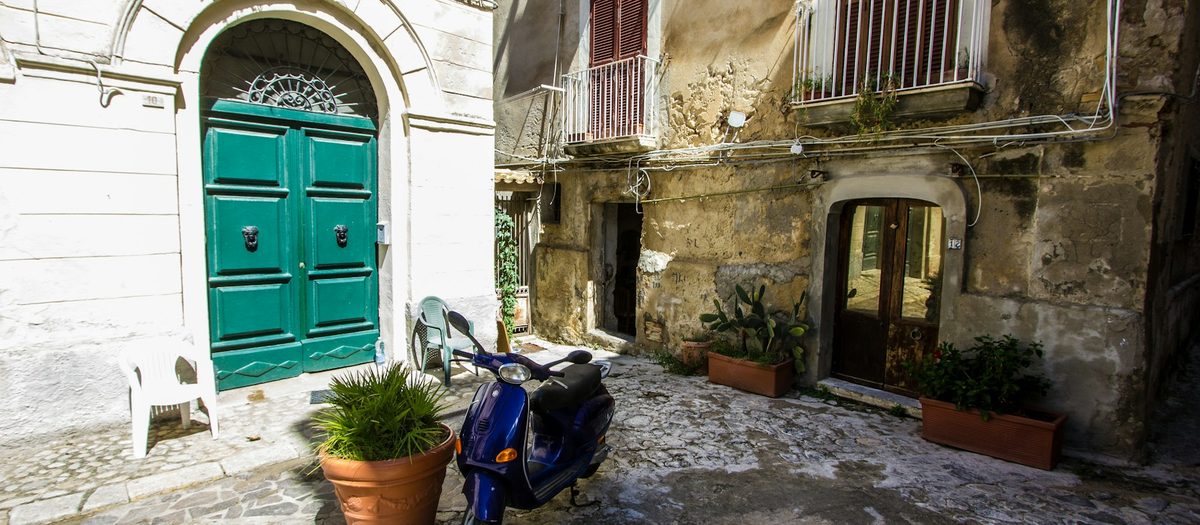 Een steegje met een groene deur, een scooter en planten in potten · Italiaanse grammatica B1