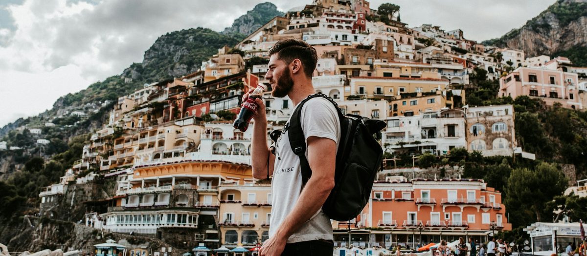 Een man met rugzak drinkt uit een fles, met Positano achter zich · Italiaanse grammatica B2