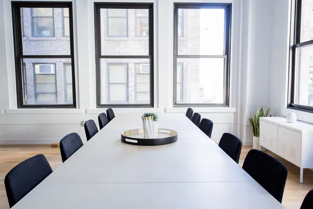 A long white meeting table with dark chairs in a bright office with tall windows