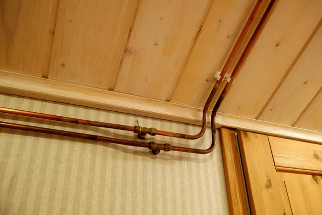 Two copper water pipes running along a wall under a wooden ceiling