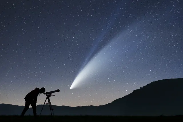 A comet with a long pale tail above a dark hillside, and an astronomer silhouetted beside a telescope