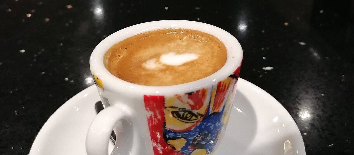 Una tazzina di caffè espresso con il piattino e il cucchiaino, sul bancone scuro di un bar.
