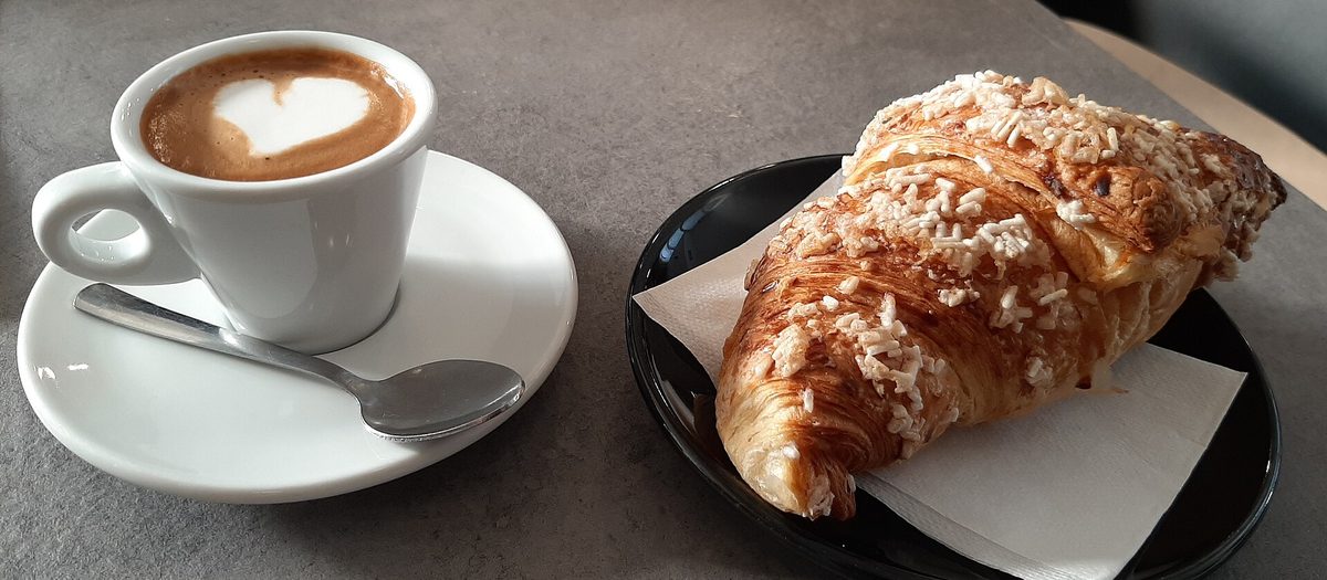 Un cappuccino con il cuore disegnato nella schiuma e un cornetto sul bancone di un bar.