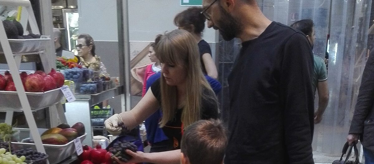 Il banco della frutta di un mercato coperto: uva, pesche e melograni nelle cassette, e la gente che compra.