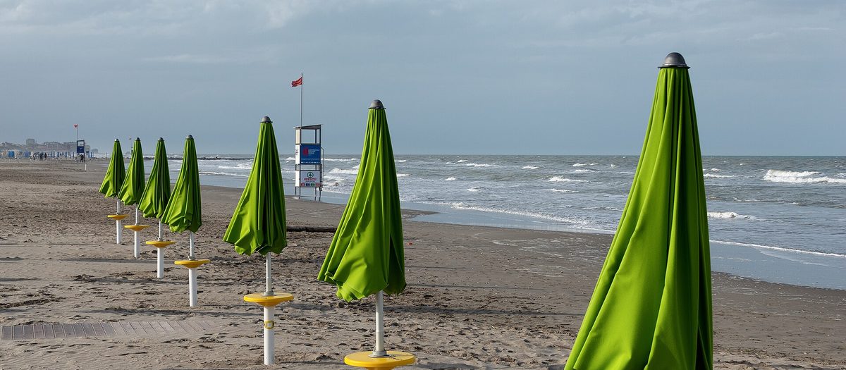 Una fila di ombrelloni verdi chiusi sulla sabbia, con il mare e la torretta del bagnino sullo sfondo.