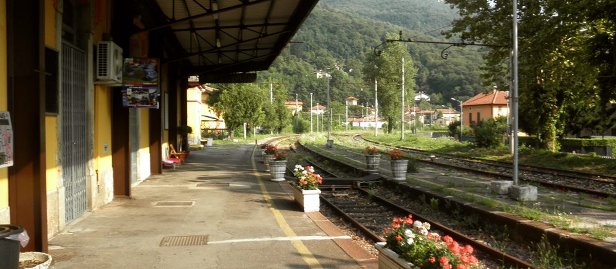 La banchina coperta di una piccola stazione di paese, con i vasi di fiori accanto ai binari e la montagna dietro.