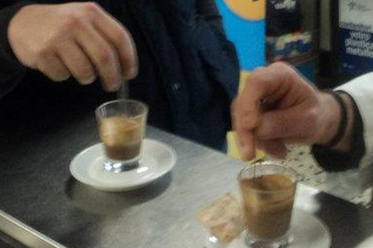 Two people stirring espressos served in small glasses on the steel counter of a bar in Rome