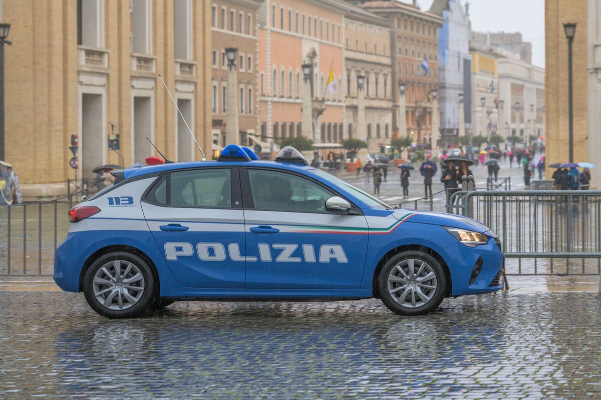 A blue and white Polizia di Stato car on the cobbles of St Peter's Square, with tourists holding umbrellas