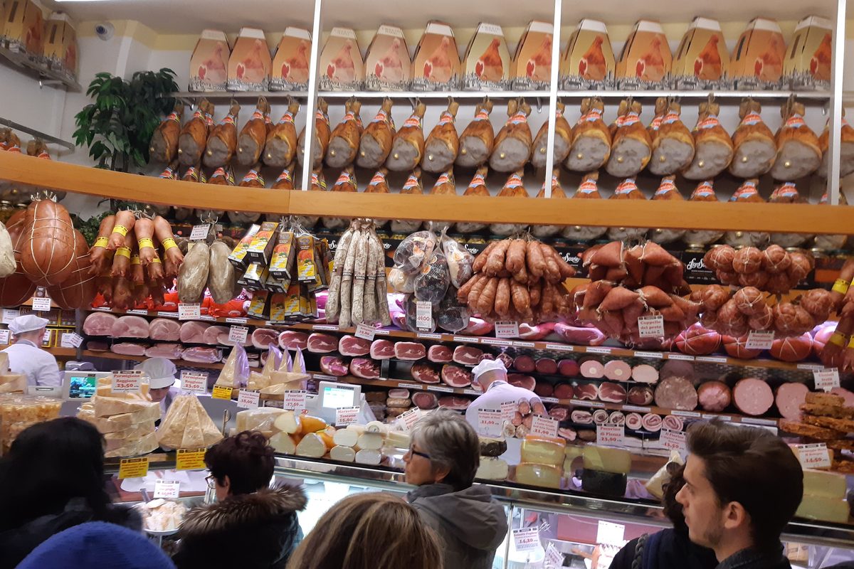 Customers at the counter of a delicatessen in Bologna, with hams and mortadella hanging overhead