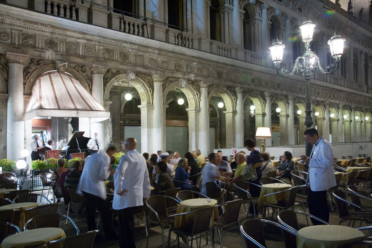 Waiters in white jackets serving full tables at a café under the arcades of St Mark's Square, Venice