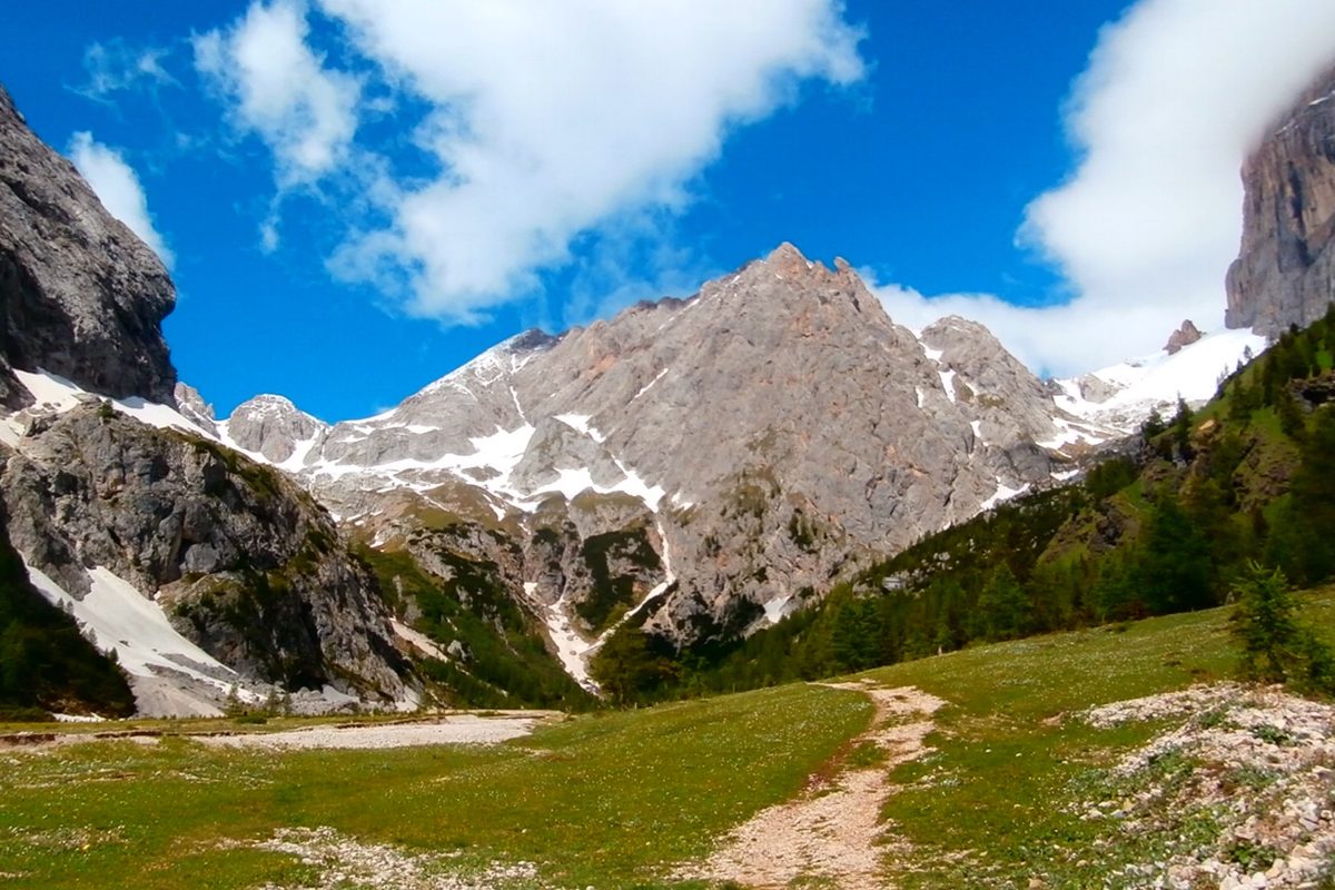 A path climbing a green valley towards the peaks of the Marmolada group