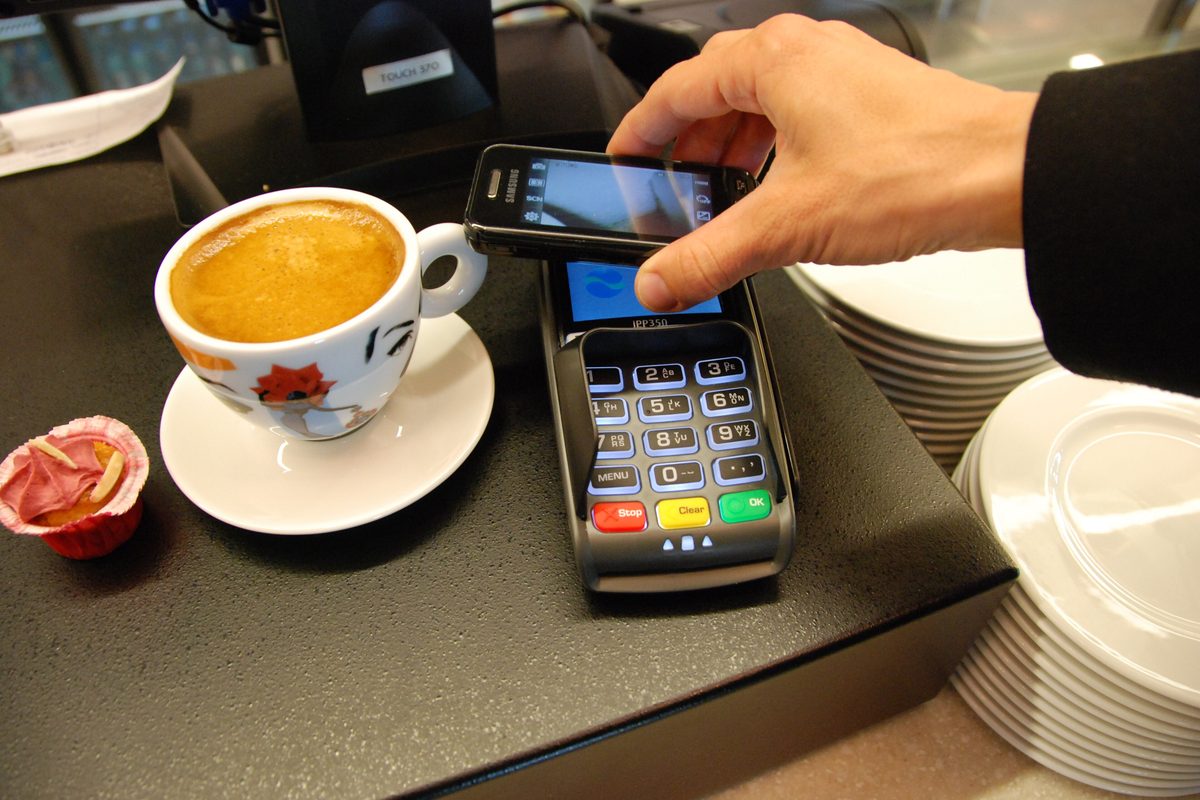 A phone held over a card terminal to pay, next to a cup of coffee on a counter