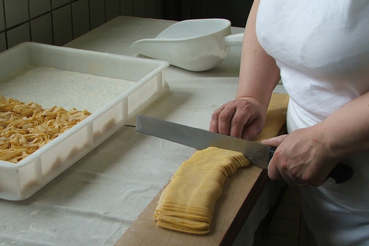 Hands cutting a rolled sheet of fresh pasta into tagliatelle with a large knife